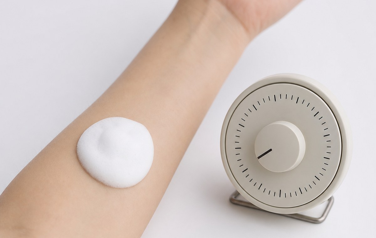 A patch of white cosmetic foam on a forearm beside a kitchen timer
